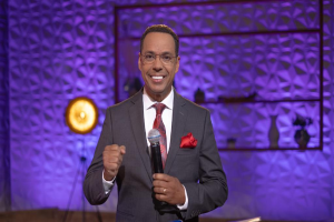 Stephen Colbert in Anzug, Krawatte und Brille steht auf einer Bühne und hält ein Mikrofon in der Hand, mit einer Wand, einem Ständer, einer Lampe und verschiedenen Gegenständen im Hintergrund.