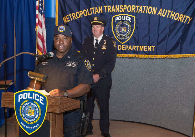 Zwei Polizeibeamte stehen vor einem Rednerpult, einer hält Papiere und spricht in ein Mikrofon, mit einem Banner, einer Fahne, einem Tisch in der Nähe und einer Tafel mit Symbolen und Text im Hintergrund.