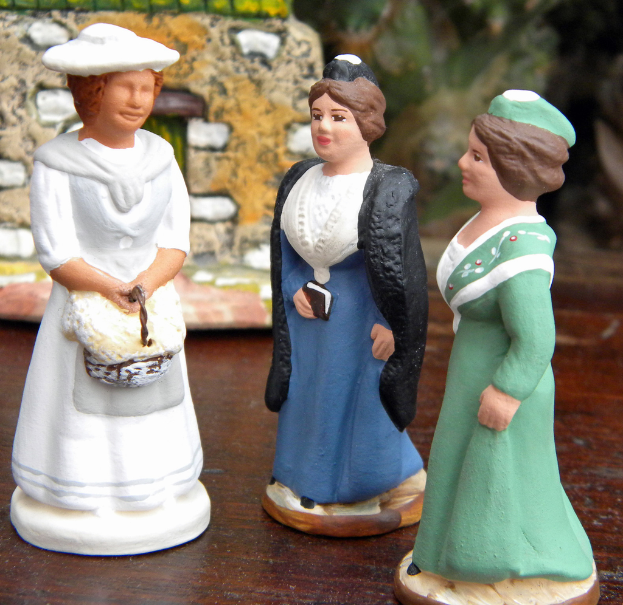 Drei kunstvoll geformte Puppen sind auf einem Holzbrett mit Spielzeughäusern im Hintergrund angeordnet.