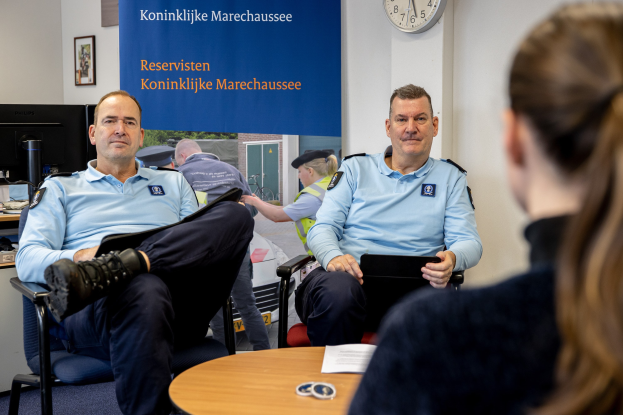 Zwei Polizeibeamte sitzen an einem Tisch mit einer Frau, umgeben von Dokumenten und Gegenständen, mit einem wandmontierten Banner, einer Uhr und Fotorahmen im Hintergrund und einem weiteren Tisch mit einem Monitor und Zubehör links.