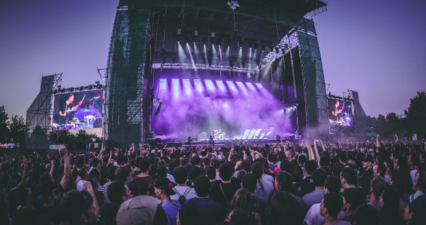 Eine große Menschenmenge bei einem Musikfestival schaut auf einer hell erleuchteten Bühne mit Instrumenten und Bildschirmen, die vor einem Gebäude, Bäumen und einem klaren blauen Himmel aufgebaut sind.