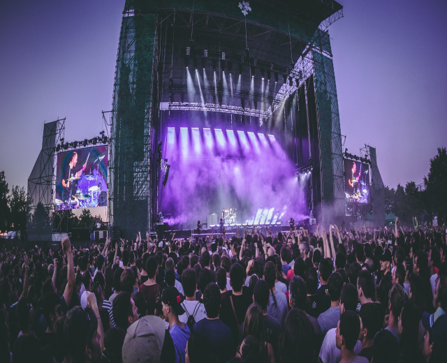 Eine große Menschenmenge bei einem Musikfestival schaut auf einer hell erleuchteten Bühne mit Instrumenten und Bildschirmen, die vor einem Gebäude, Bäumen und einem klaren blauen Himmel aufgebaut sind.