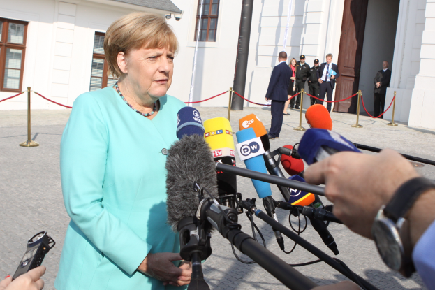 Bundeskanzlerin Angela Merkel spricht vor einem Gebäude mit Fenstern und einer Tür zu den Medien, umgeben von einer Gruppe von Menschen, wobei auf der rechten Seite eine Kamera und ein Mobiltelefon sowie Pfosten mit daran gebundenen Seilen zu sehen sind.
