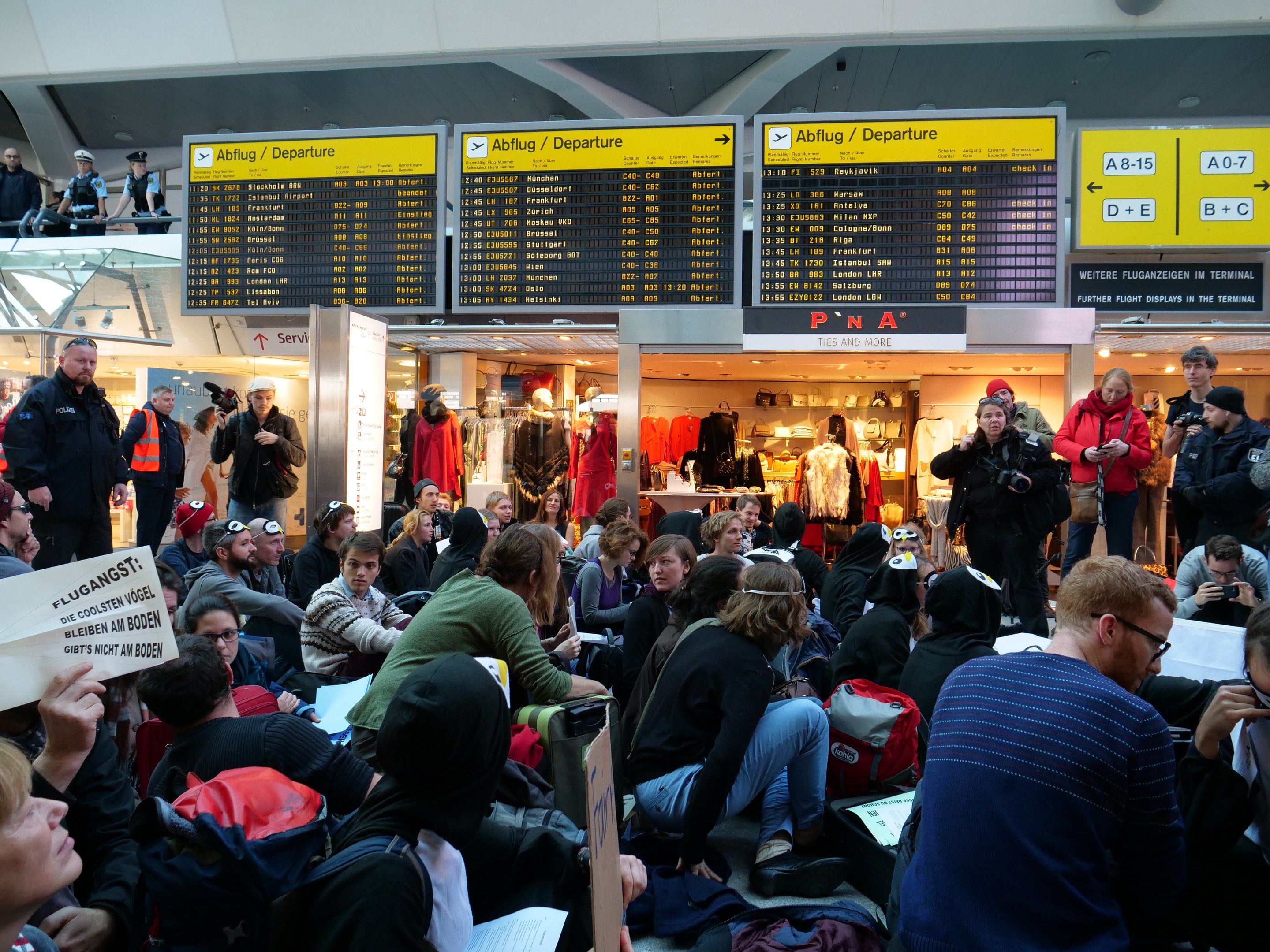 Große Gruppe von Menschen an einem Flughafen, einige sitzen mit Taschen und Papieren, andere stehen, mit Texttafeln, Schaufensterpuppen in Kleidern und Deckenbeleuchtung im Hintergrund, was auf eine Demonstration hinweist.
