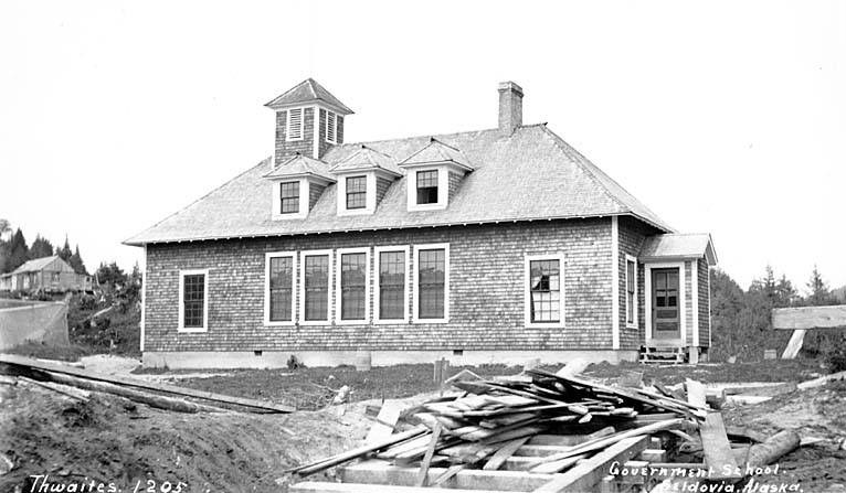 Schwarz-weiß-Foto einer Regierungsschule aus dem Jahr 1905, umgeben von Bäumen unter einem klaren Himmel, mit Holzplanken und der Beschriftung "Regierungsschule, Thwaites, 1905" unten.