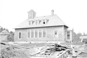 Schwarz-weiß-Foto einer Regierungsschule aus dem Jahr 1905, umgeben von Bäumen unter einem klaren Himmel, mit Holzplanken und der Beschriftung "Regierungsschule, Thwaites, 1905" unten.