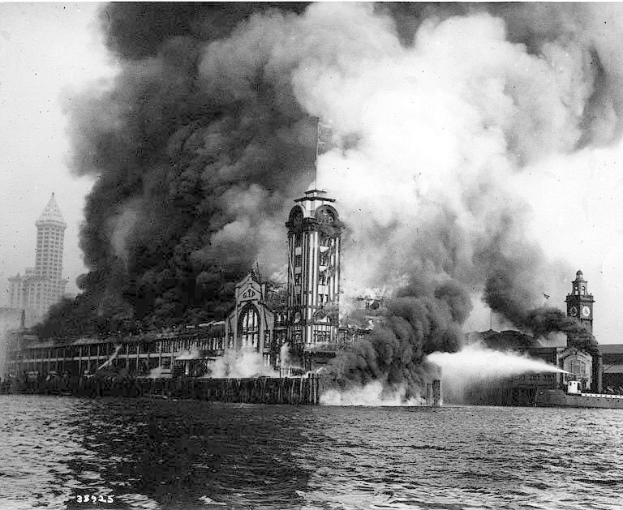 Schwarz-weiß-Foto eines großen, von Flammen umloderten Gebäudes mit Rauchwolken, einem Gewässer im Vordergrund und anderen Gebäuden und Himmel im Hintergrund.