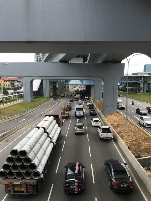 Eine Autobahn mit starkem Verkehr unter einer Brücke, umgeben von Masten, Straßenlaternen, Gras, Gebäuden, Bäumen und dem Himmel.