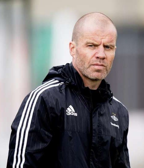 Ein kahlköpfiger Mann in einer schwarzen Jacke, der wie ein Trainer aussieht, steht auf einem Fußballfeld mit ernster Miene, trägt eine blaue Jacke mit weißen Streifen und einem weißen Kragen und hat die Hände vor der Brust verschränkt.