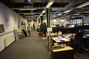 Ein geräumiges Großraumbüro mit zahlreichen Schreibtischen, Stühlen und Computern, Menschen bei der Arbeit, verschiedenen Gegenständen auf den Tischen, Kleidung an der Garderobe, Wanddisplays und Deckenleuchten, mit Glasfenstern im Hintergrund.