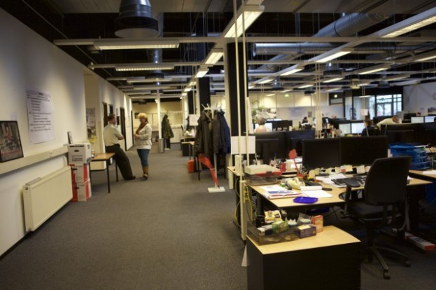 Ein geräumiges Großraumbüro mit zahlreichen Schreibtischen, Stühlen und Computern, Menschen bei der Arbeit, verschiedenen Gegenständen auf den Tischen, Kleidung an der Garderobe, Wanddisplays und Deckenleuchten, mit Glasfenstern im Hintergrund.