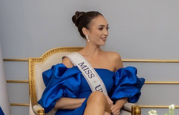 Frau in einem blauen Kleid mit einem "Miss Universe 2019"-Band, das auf einem Stuhl sitzt und von Blumen umgeben ist, mit einer Wand im Hintergrund.