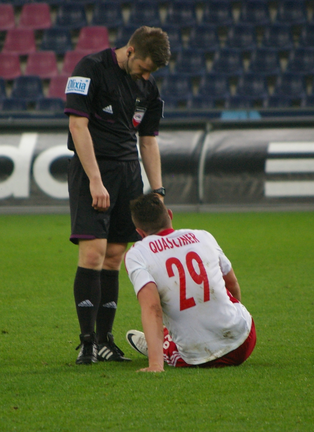 Ein Fußballspieler sitzt neben einem Schiedsrichter auf dem Boden in einem Stadion, beide tragen Sportkleidung, mit Brettern und Stühlen im Hintergrund.