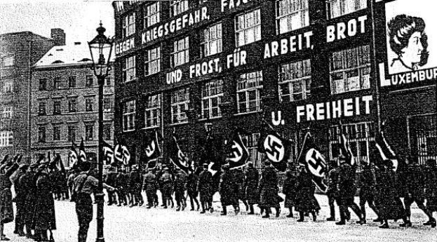 Schwarzes und wei├čes Bild einer Gruppe, die mit Fahnen und Transparenten die Stra├če entlangmarschiert, mit einem Lichtmast links und einem Gebäude mit einem Nazi-Parteizeichen im Hintergrund.