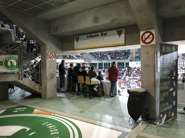 Eine Gruppe von Menschen steht vor einer sitzenden Menge in einem Stadion während eines Boston Celtics vs New York Jets-Events, mit Schildern, einem Müllcontainer, einer Texttafel, einer Treppe und Deckenleuchten.