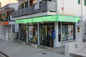 Apotheke an einer Straßenecke mit Gebäuden, Geschäften, Schildern, Strommasten, Kabeln, Fahrzeugen, Topfpflanzen und einem klaren blauen Himmel.