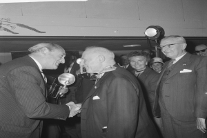 Zwei Männer schütteln sich die Hand vor einem lachenden Publikum, einer hält ein Mikrofon, unter einem Licht, dargestellt in Schwarz-Weiß.