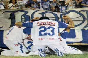 Gruppe von Fußballspielern, die auf dem Boden vor einer jubelnden Menge feiern, mit einem Banner im Hintergrund.