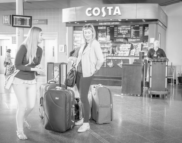 Zwei Frauen mit Gepäck vor sich, mit einem Kaffee-Shop im Hintergrund, wahrscheinlich an einem Flughafen.