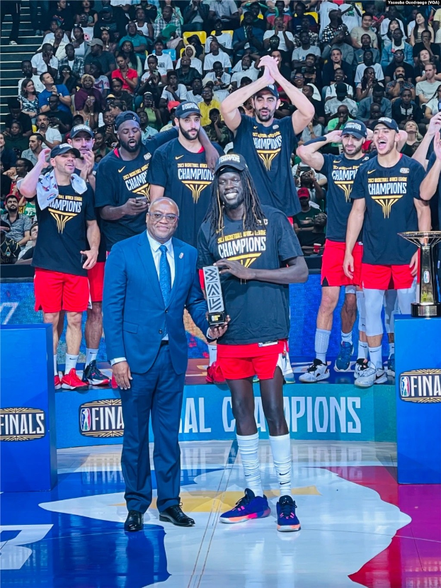 Eine Gruppe von Männern auf einem Basketballfeld, die Trophäen halten, mit Zuschauern im Hintergrund und zwei Trophäen auf Podesten, was einen Sieg bei den NBA-Finals suggeriert.