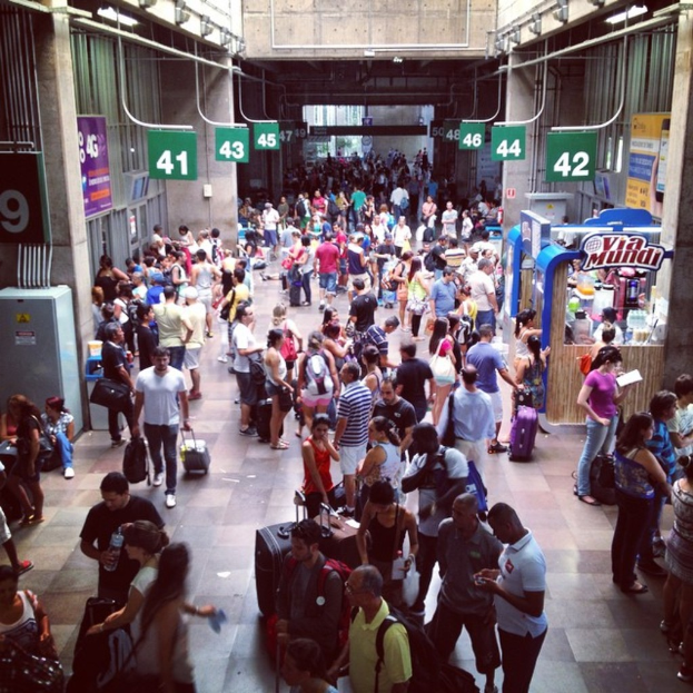Eine Gruppe von Menschen auf einem Weg, einige tragen Gepäck, mit Ständen, Kisten, Rohren, Brettern, Schildern, Wänden und verschiedenen Gegenständen drumherum.