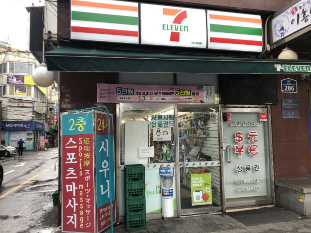 Ein 7-Eleven-Konzern in Seoul, Südkorea, mit Fahrzeugen auf der Straße, einem Fußgänger auf der linken Seite und Gebäuden im Hintergrund unter einem sichtbaren Himmel.