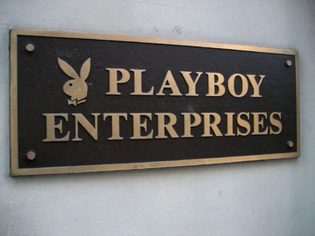 Ein rechteckiges Namensschild an einer Wand mit sichtbaren Schrauben, das ein Logo und Text enthält.