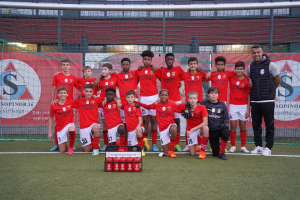 Eine Gruppe junger Männer in roten T-Shirts und schwarzen Shorts steht auf einem Fußballfeld vor einer Kiste roter Gegenstände, mit Bannern im Hintergrund, die "FC Red Black Eglaute - Champions League" lesen.