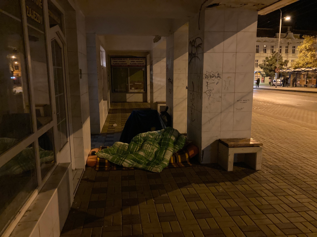 Eine Person schläft auf einer Decke und einer Bank auf einem Gehweg bei Nacht, mit einer Gruppe von Menschen in der Nähe, Fahrzeugen auf der Straße, einem Straßenpfahl, Bäumen, einem Schild, Gebäuden mit Fenstern und einem bewölkten Himmel im Hintergrund.
