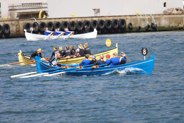 Eine Gruppe von Menschen in einem blauen und gelben Boot auf dem Wasser, die Paddel halten, mit einer Wand mit Reifen und einem Gebäude im Hintergrund.