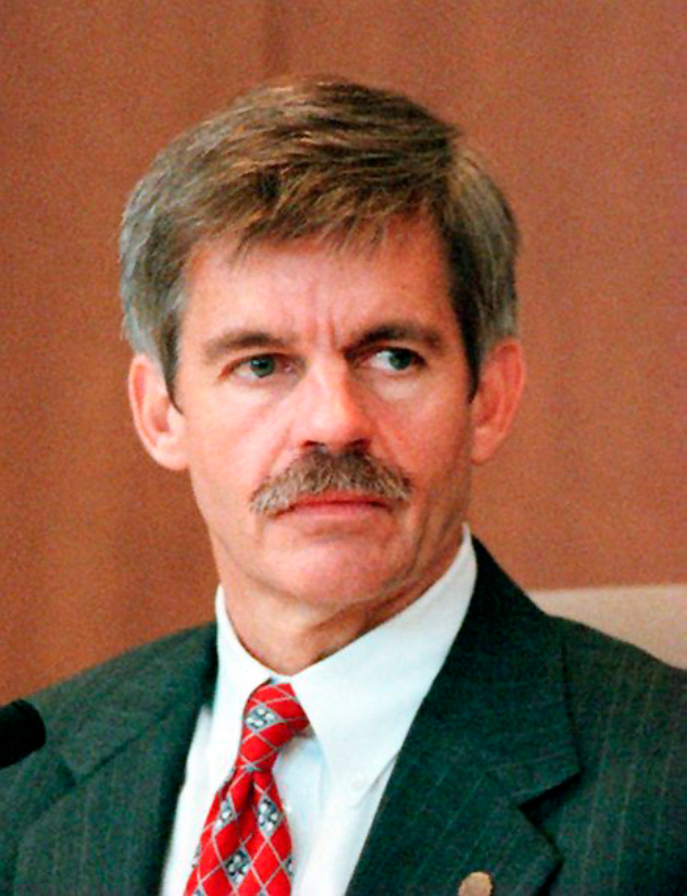 Ein Mann in einem marineblauen Anzug, weißem Hemd und roter Krawatte sitzt an einem Tisch mit einem Mikrofon, sieht ernst aus mit ordentlich gekämmtem Haar, vor einer Wand im Hintergrund.