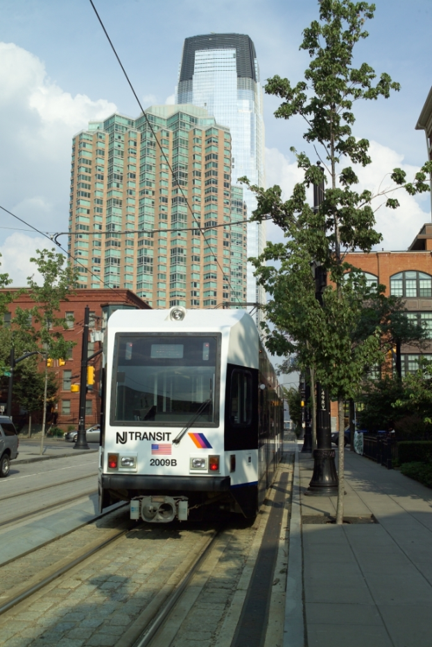 Ein Zug auf Schienen mit elektrischen Kabeln darauf, umgeben von Bäumen, Masten und Gebäuden, mit einem Wolkenkratzer und Himmel im Hintergrund.