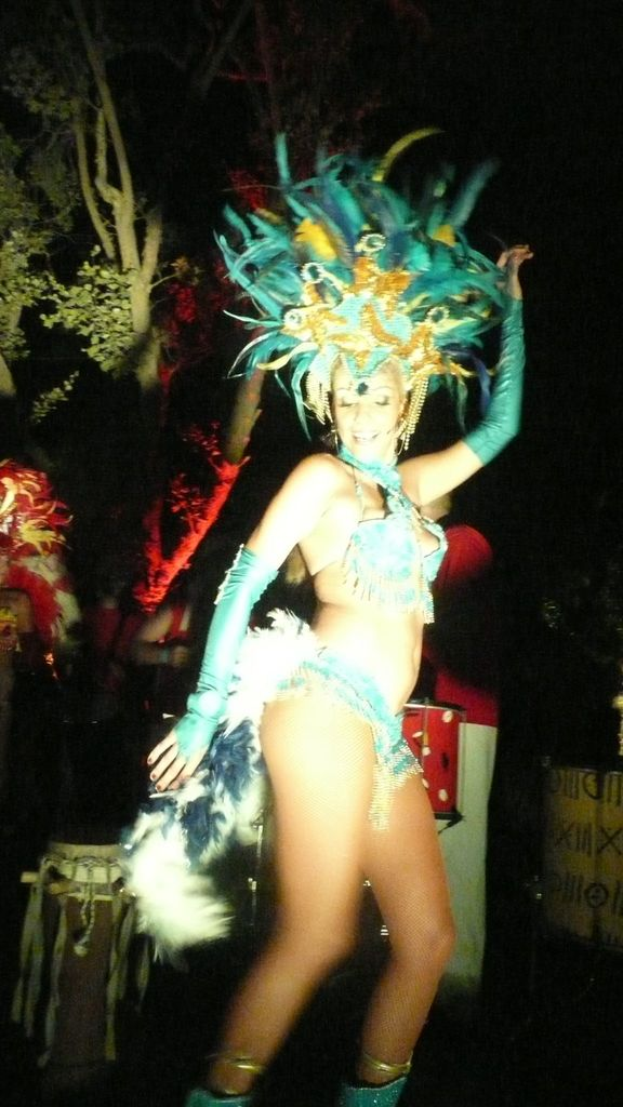 Eine Frau in einem eleganten Kostüm tanzt mit Trommeln und anderen Menschen im Hintergrund, mit einem Baum in der Nähe.