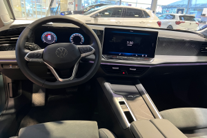 Das Innere eines Volkswagen e-Golf, mit einem Lenkrad, Armaturenbrett, Bildschirmen, Knöpfen, Sitzen und Glastüren, mit Blick auf andere Autos, Gebäude, Bäume, Pfosten und den Himmel.