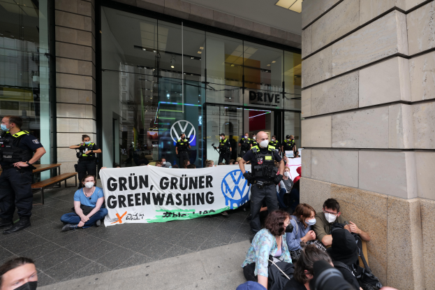 Eine Gruppe von Menschen sitzt vor einer Volkswagen-Niederlassung auf dem Boden und hält ein Transparent mit Text, einige tragen Masken und andere halten Kameras, im Hintergrund sind Bänke und ein Gebäude mit Glaswänden zu sehen, sowie Lampen an der Decke.