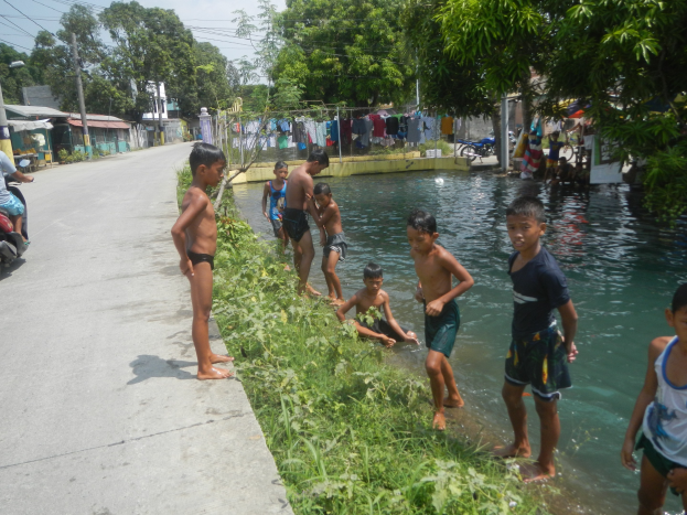 Kinder spielen in flachem Wasser neben einer Straße, einige stehen und andere sitzen auf Gras, mit einer Person auf einem Motorrad links und Häusern, Bäumen und Strommasten im Hintergrund unter einem klaren blauen Himmel.