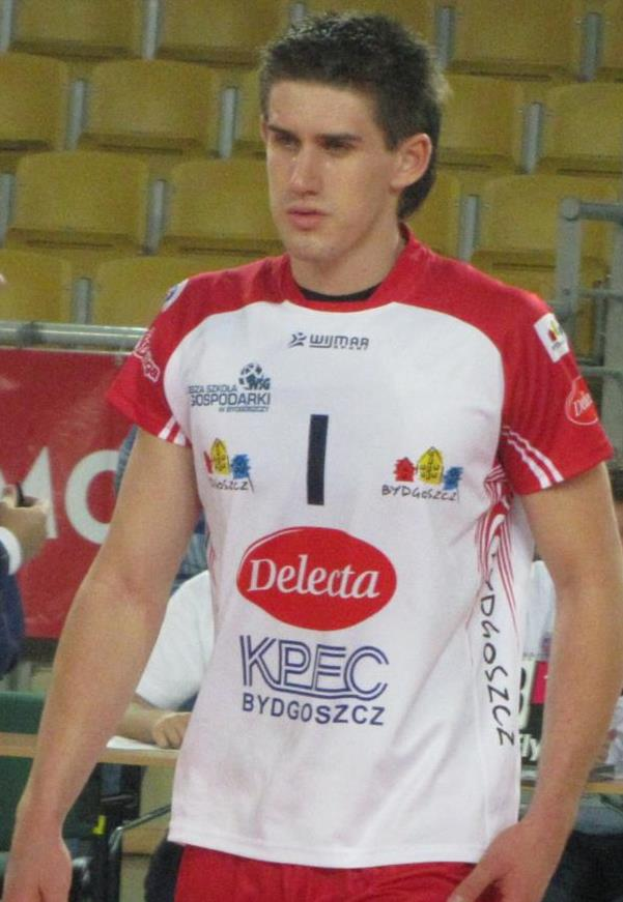 Ein Mann in einem roten und weißen Handballtrikot steht auf einem Platz, umgeben von Zuschauern, mit einem roten Banner im Hintergrund, auf dem "Krasnoyarsk vs Bydgoszcz" steht.