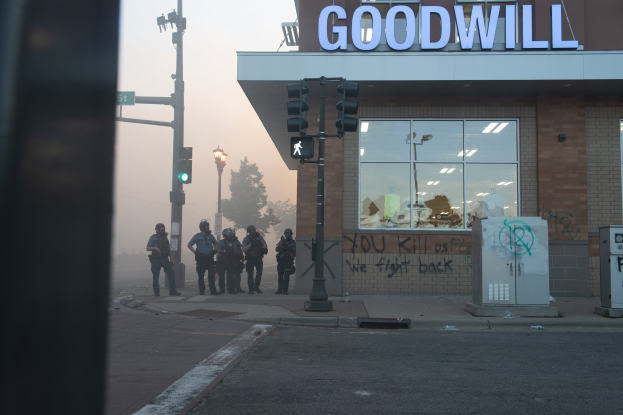 Eine Gruppe von Polizisten in Helmen und taktischer Ausrüstung steht vor einem Glasfront-Gebrauchtwarenladen, bewaffnet, mit Verkehrssignalen und Laternen im Vordergrund und Bäumen unter einem klaren Himmel im Hintergrund.