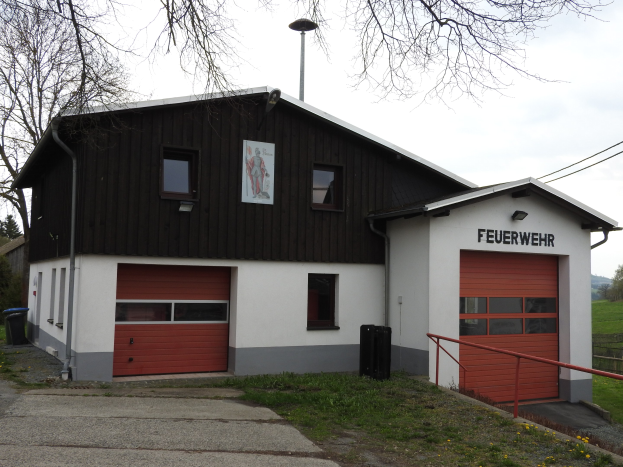 Rote und weiße Feuerwache mit einer Straße, Gras, gelben Blumen und Bäumen im Vordergrund und dem Himmel im Hintergrund.