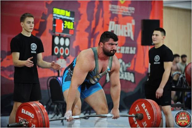 Ein Mann hebt eine Hantel bei einem Gewichtheben-Wettkampf, umgeben von Zuschauern, mit Wettkampfausrüstung und Beschilderung, die die 2018 World Weightlifting Championships anzeigt.