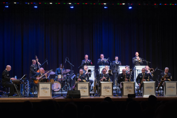 Eine Gruppe von Musikern tritt auf einer hell erleuchteten Bühne auf, die mit "The New Orleans Jazz Band" beschriftet ist, während Mikrofone, Ständer und Texttafeln im Hintergrund zu sehen sind und das Publikum darunter zusieht.