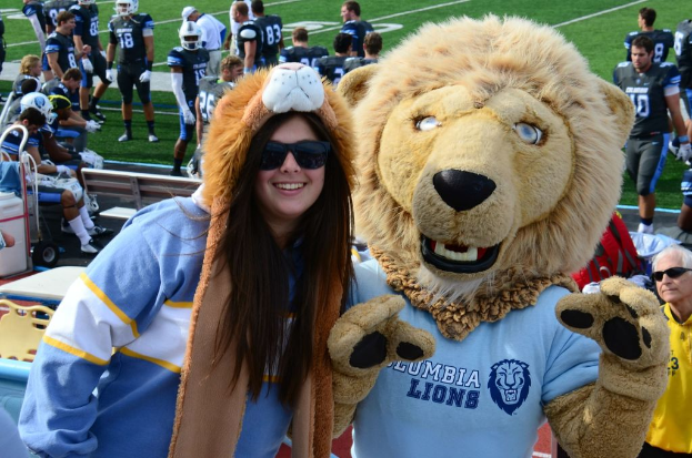 Eine Frau steht in der Nähe einer Person in einem Löwenkostüm, mit Spielern auf dem Feld und Zuschauern auf Bänken und Stühlen im Hintergrund.