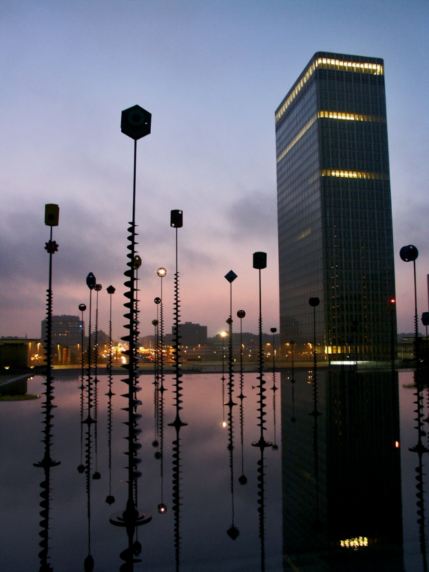 Mehrere Masten und Gebäude mit Lichtern unter einem bewölkten Himmel, mit reflektierenden Masten.