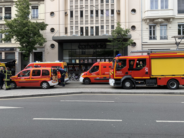 Eine Gruppe von Feuerwehrautos auf einer Straße in Paris geparkt, mit Menschen auf dem Gehweg in der Nähe, Gebäuden, Bäumen und einem Fahrrad im Hintergrund.