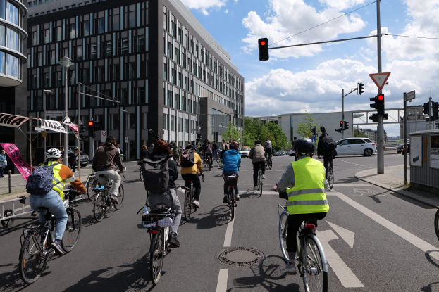 Eine Gruppe von Menschen, die auf Fahrrädern eine Straße mit hohen Gebäuden entlangfahren, wobei einige Fahrer Helme und Taschen tragen, andere auf dem Gehweg stehen, Fahrzeuge auf der Straße, Verkehrszeichen, Schilder, Bäume und ein bewölkter Himmel im Hintergrund zu sehen sind.