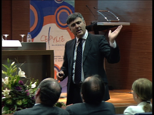 Ein Mann in einem schwarzen Blazer steht an einem Podium mit Mikrofonen, spricht in ein tragbares Mikrofon, mit einer Blumenanordnung und einem Tisch mit einem Buch und einer Wasserflasche in der Nähe, während drei Personen auf Stühlen vor ihm sitzen, vor einem Hintergrund aus einer braunen Wand und einem bunten Schild mit Text.