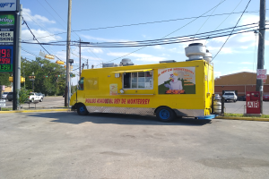 Ein gelber Foodtruck vor einer Tankstelle geparkt, umgeben von Strommasten mit Kabeln und Schildern mit Text und Zahlen, mit Bäumen, Gebäuden und einem bewölkten Himmel im Hintergrund.