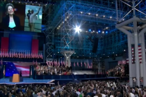 Große Menschenmenge vor einer Bühne mit Podium, Mikrofon, Flaggen, einem Overhead-Bildschirm und Lichtinstallationen bei dem, was wie eine Präsidentschaftsdebatte aussieht.