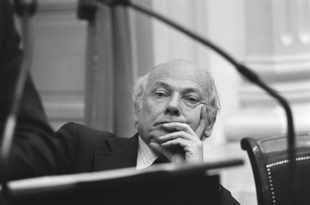 Ein Mann in einem Anzug sitzt an einem Tisch und hat seine Hand auf seinem Kinn, umgeben von einem Mikrofon und einem Buch, mit einem unscharfen Hintergrund.