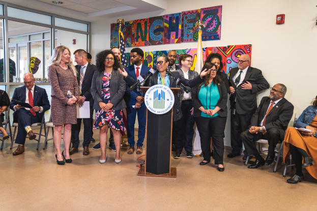 Eine Gruppe von Menschen, die in einem Raum mit einem Podium, einem Mikrofon und einer Tafel mit der Aufschrift "Sancho City Councilman Joins City Council" stehen und sitzen, mit Flaggen, Feuerl"oscher und Deckenleuchten an der R"uckwand.